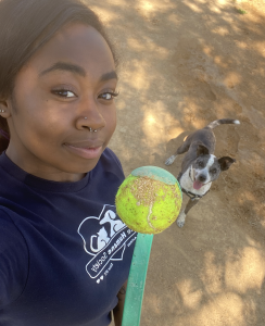 Volunteer posing for a selfie with a shelter dog in the background. She is holding up a ball and the dog looks happily at the camera